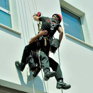 Two people in harnesses are suspended by ropes, descending a building near a window. Its an adventure camp scene in Belleville, Ontario, where one appears to assist the other. Both wear helmets and safety gear against the backdrop of the white building with its nearby window.